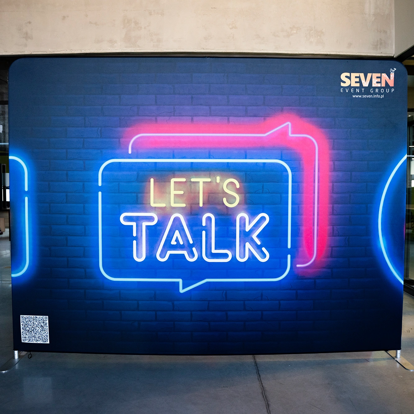 Ścianka tekstylna z nadrukiem imitującym neon „LET’S TALK” na tle ceglanej ściany, przygotowana na potrzeby eventu organizowanego przez Seven Event Group.