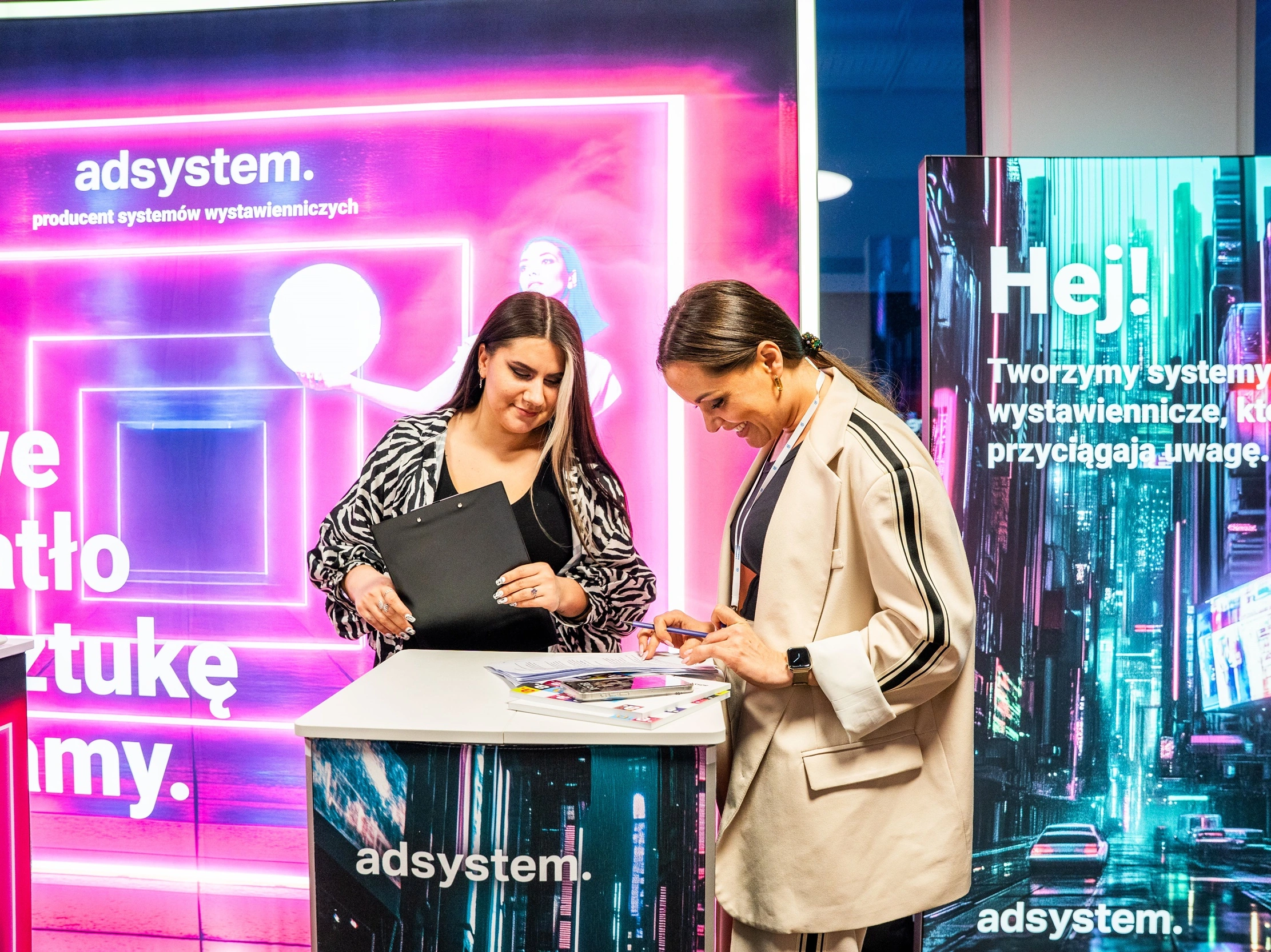 Dwie kobiety rozmawiają przy trybunce reklamowej adsystem, na tle podświetlanych systemów wystawienniczych z neonowymi grafikami i hasłami.