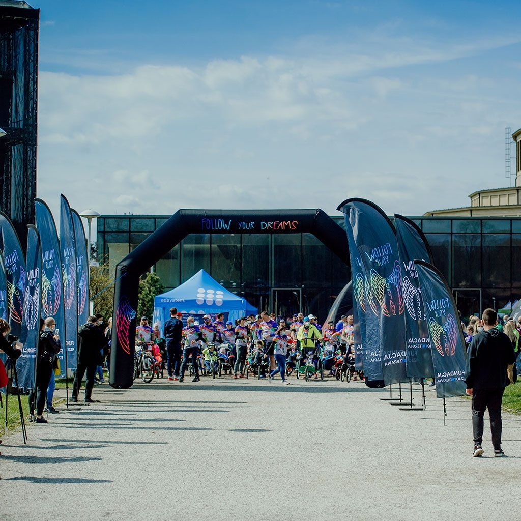 Outdoor sports event with branded advertising flags, an inflatable arch, and a blue promotional tent, with participants gathering at the starting zone.