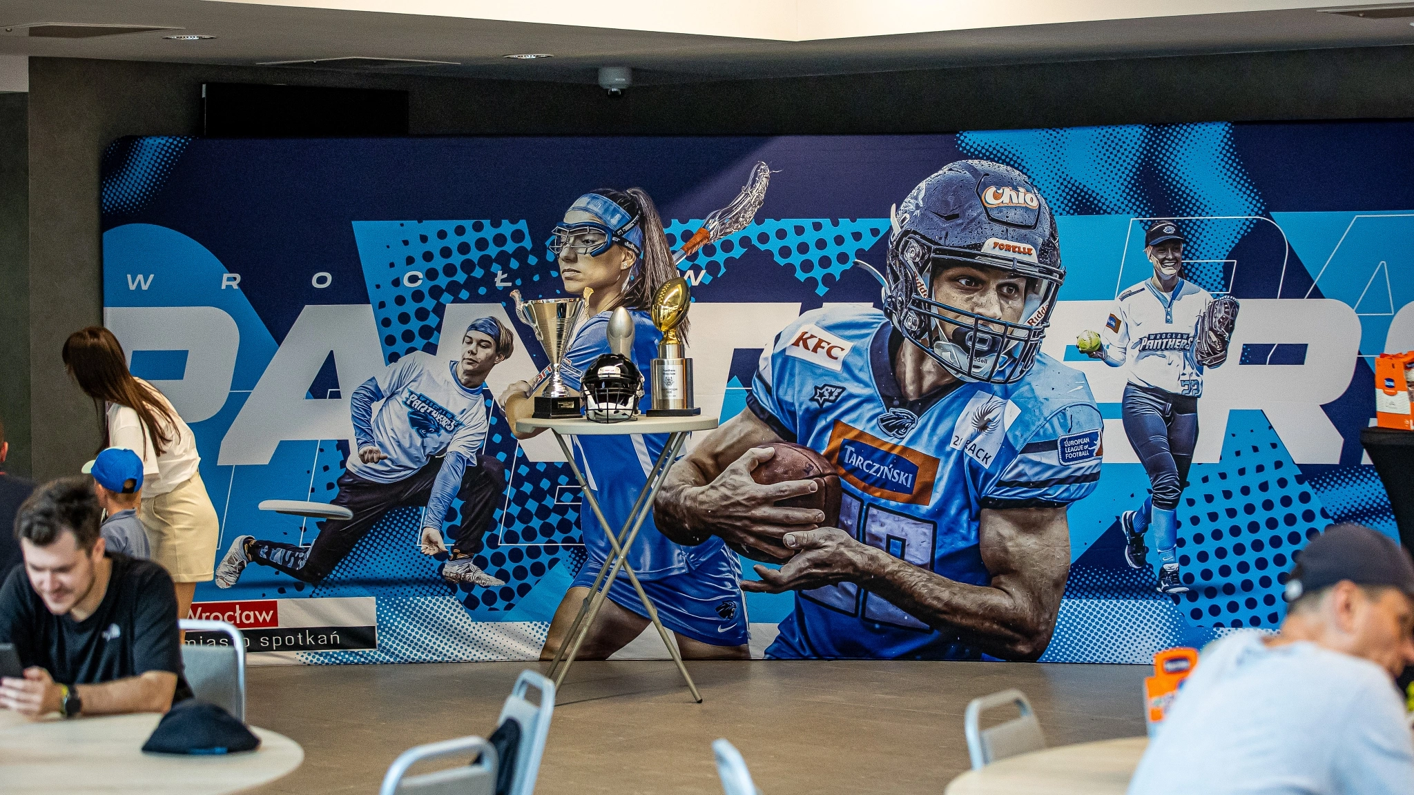 Ścianka tekstylna z grafiką promującą drużynę Panthers Wrocław, prezentująca zawodników różnych dyscyplin i ustawiona w strefie eventowej z pucharami i gadżetami klubowymi.
