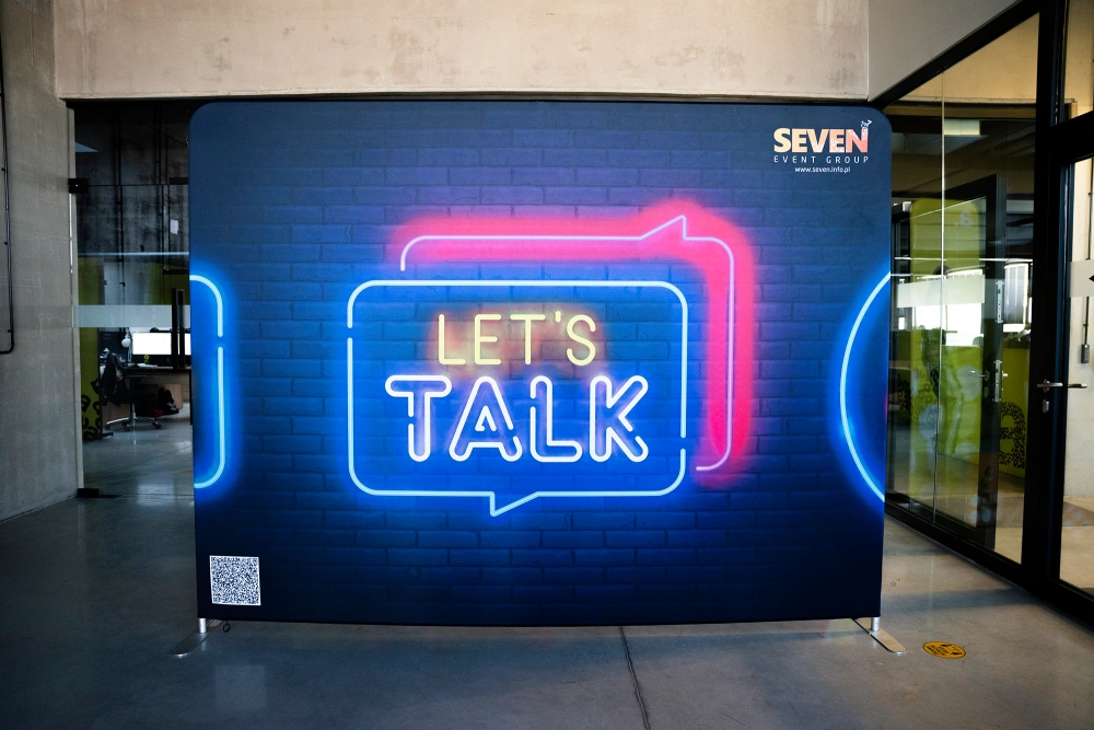 Photocall textil publicitario para ferias con estructura modular de aluminio y gráfica impresa doble cara con diseño de neón “Let’s Talk”.