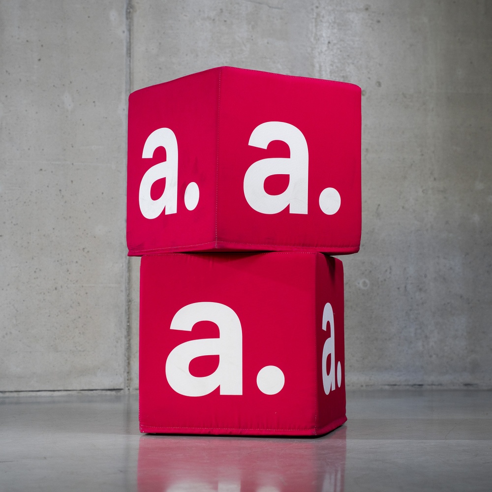 Dwie pufy kostki z wymiennymi, czerwonymi pokrowcami z nadrukiem „a.”, gotowe do ponownego wykorzystania w różnych aranżacjach.