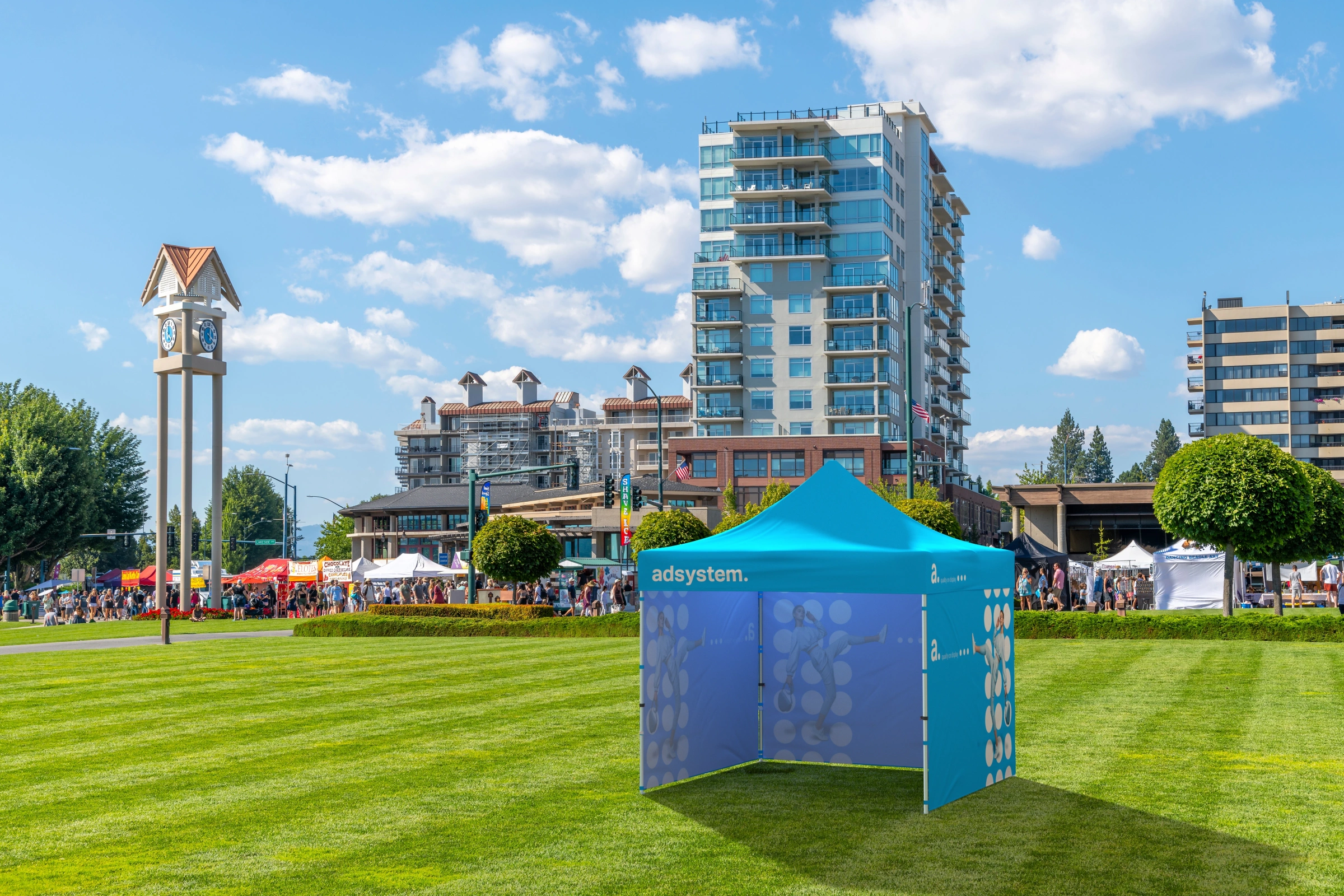 Niebieski namiot reklamowy adtent express 3x3 ustawiony na zielonym trawniku w centrum miasta, w tle nowoczesne budynki, wieża zegarowa i tłum uczestników wydarzenia plenerowego