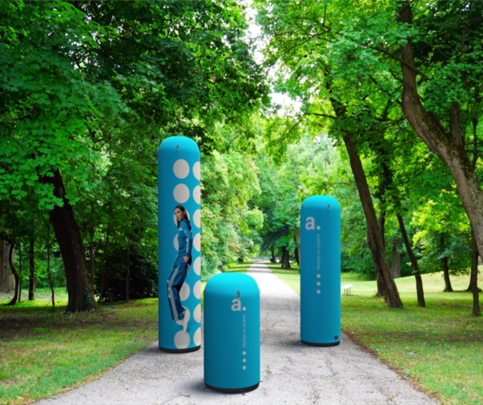 Postes publicitarios hinchables de diferentes tamaños con diseño azul y elementos gráficos, colocados en un camino dentro de un parque arbolado.