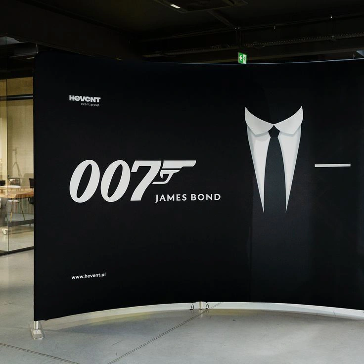 Czarna ścianka tekstylna z motywem Jamesa Bonda, ozdobiona logotypem „007” z ikoną pistoletu i grafiką smokingu, ustawiona w eleganckim wnętrzu eventowym.
