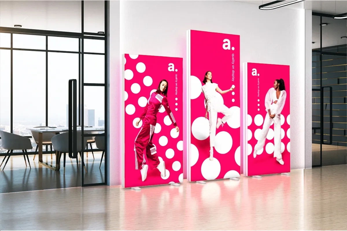 Trzy podświetlane kasetony reklamowe adFrame ustawione w nowoczesnym biurze; każdy z nich prezentuje dynamiczne zdjęcie kobiety na tle różowej grafiki z białymi kropkami, podkreślając nowoczesny design i wyrazistą identyfikację wizualną.