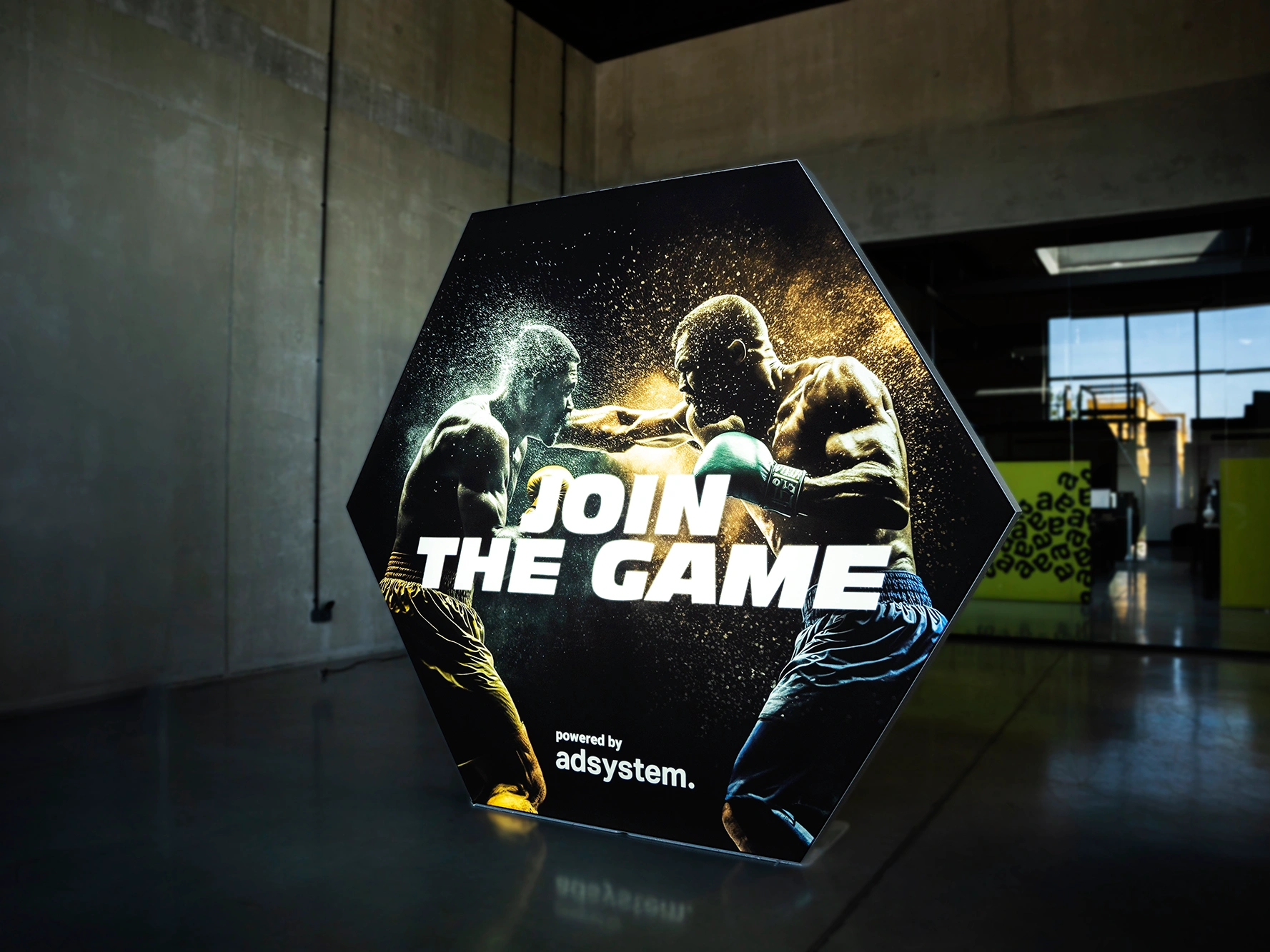 Kaseton w kształcie sześciokąta, podświetlana grafika z motywem bokserskim, dynamiczna scena pojedynku, hasło „Join the Game”, oznaczenie „powered by adsystem”.