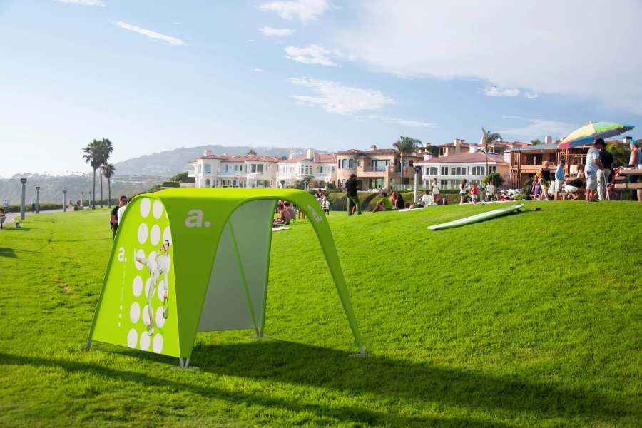 Lekki namiot reklamowy z grafiką , rozstawiony na zielonym trawniku w parku wśród ludzi i domów w tle.