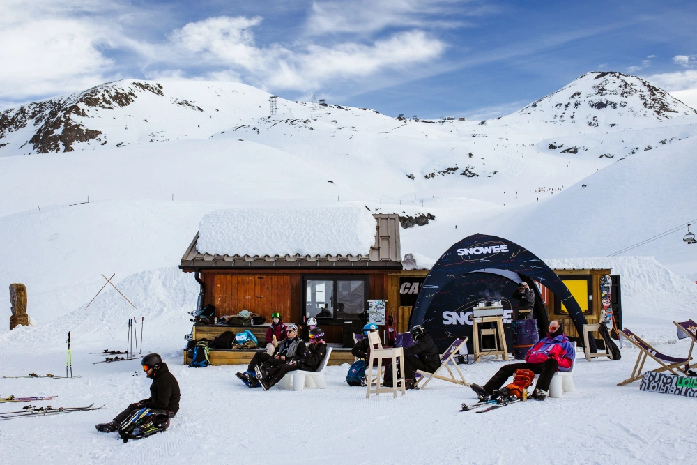 Strefa marki Snowee podczas Snowee Music Trip 2024 w Alpach – namiot reklamowy adsystem, flagi i leżaki na tle ośnieżonych gór i narciarzy odpoczywających po zawodach.