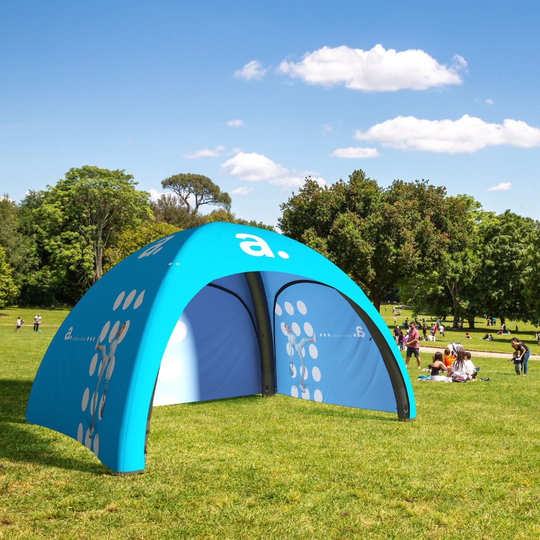 Niebieski namiot reklamowy ustawiony w zielonym parku, w tle grupy ludzi odpoczywających na trawie pod drzewami w słoneczny dzień.