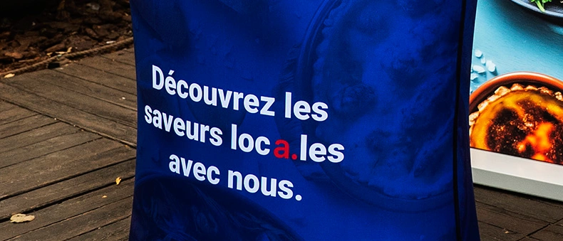 Zbliżenie na trybunkę reklamową z nadrukiem „Découvrez les saveurs locales avec nous” w niebieskiej oprawie.