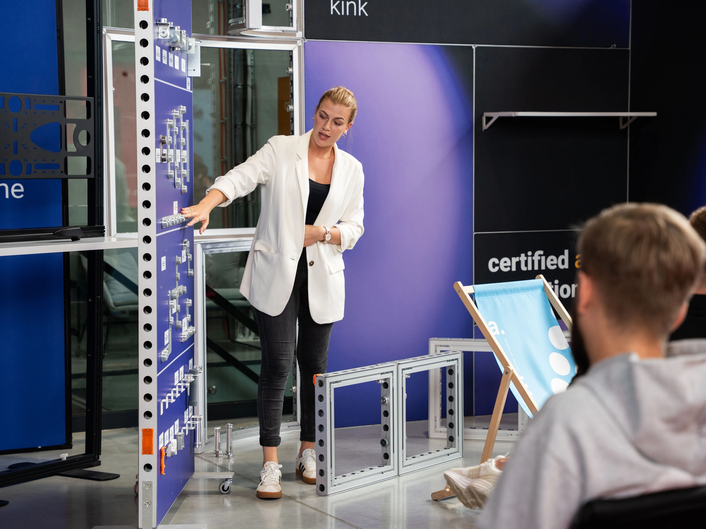 Kobieta w białej marynarce prezentuje elementy systemu wystawienniczego przed grupą słuchaczy.