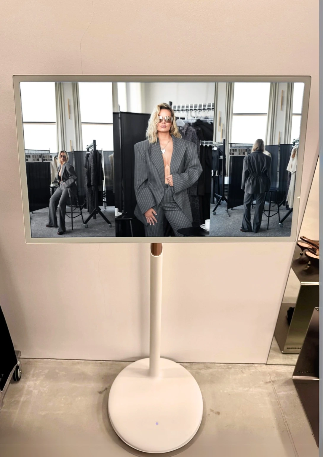  Infokiosk adVideo Stand 32’ w showroomie modowym, wyświetlający zdjęcia modelki w szarym garniturze w prążki. Nowoczesny ekran na białej nodze prezentuje treści wizualne w eleganckim otoczeniu wnętrza fashion.