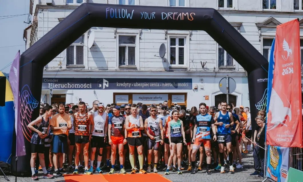 A group of runners lined up at the starting line under a large inflatable arch that reads 'FOLLOW YOUR DREAMS'. The event is taking place on a sunny day with a visible backdrop of a city building and banners displaying logos.