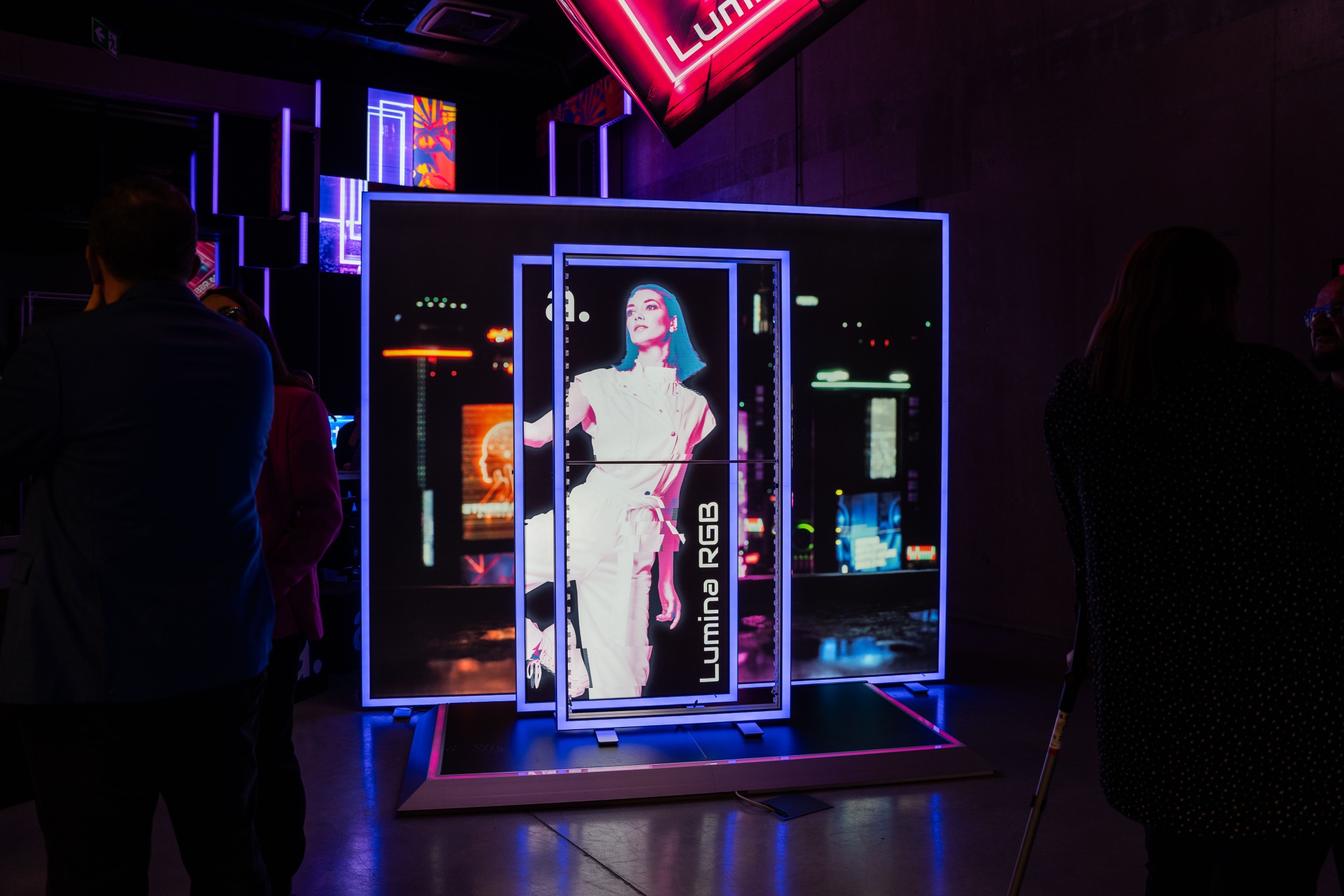 Ekspozycja systemu reklamowego adFrame Lumina RGB w ciemnym, nowoczesnym wnętrzu, z podświetlaną grafiką i elementami neonowymi, przedstawiająca kobietę w jasnym stroju na tle miejskich świateł, otoczona oglądającymi uczestnikami wydarzenia.