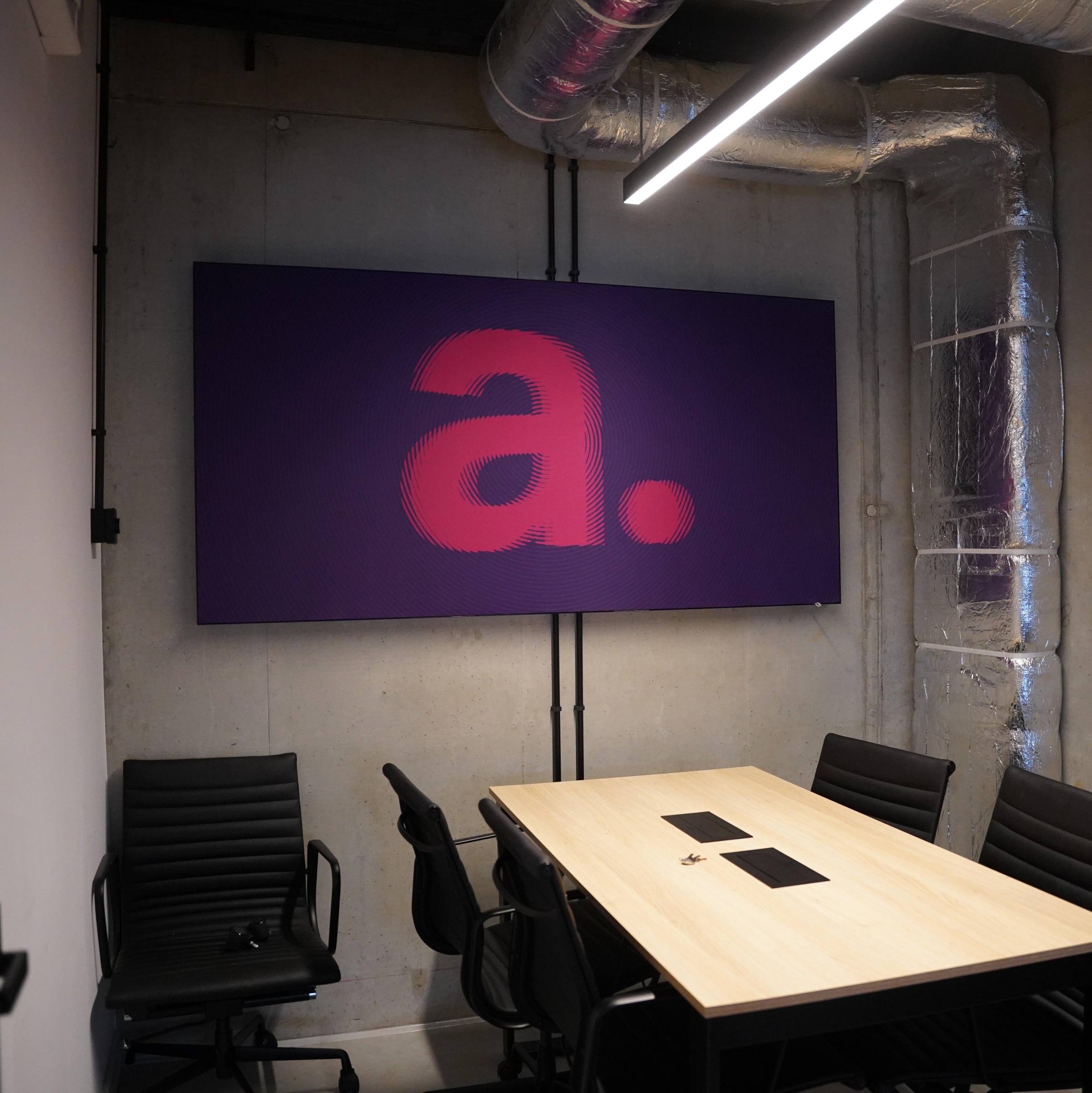 Nowoczesna sala konferencyjna z drewnianym stołem, czarnymi krzesłami i dużym fioletowym panelem z różowym logotypem „a.” na betonowej ścianie, z widocznymi instalacjami wentylacyjnymi.