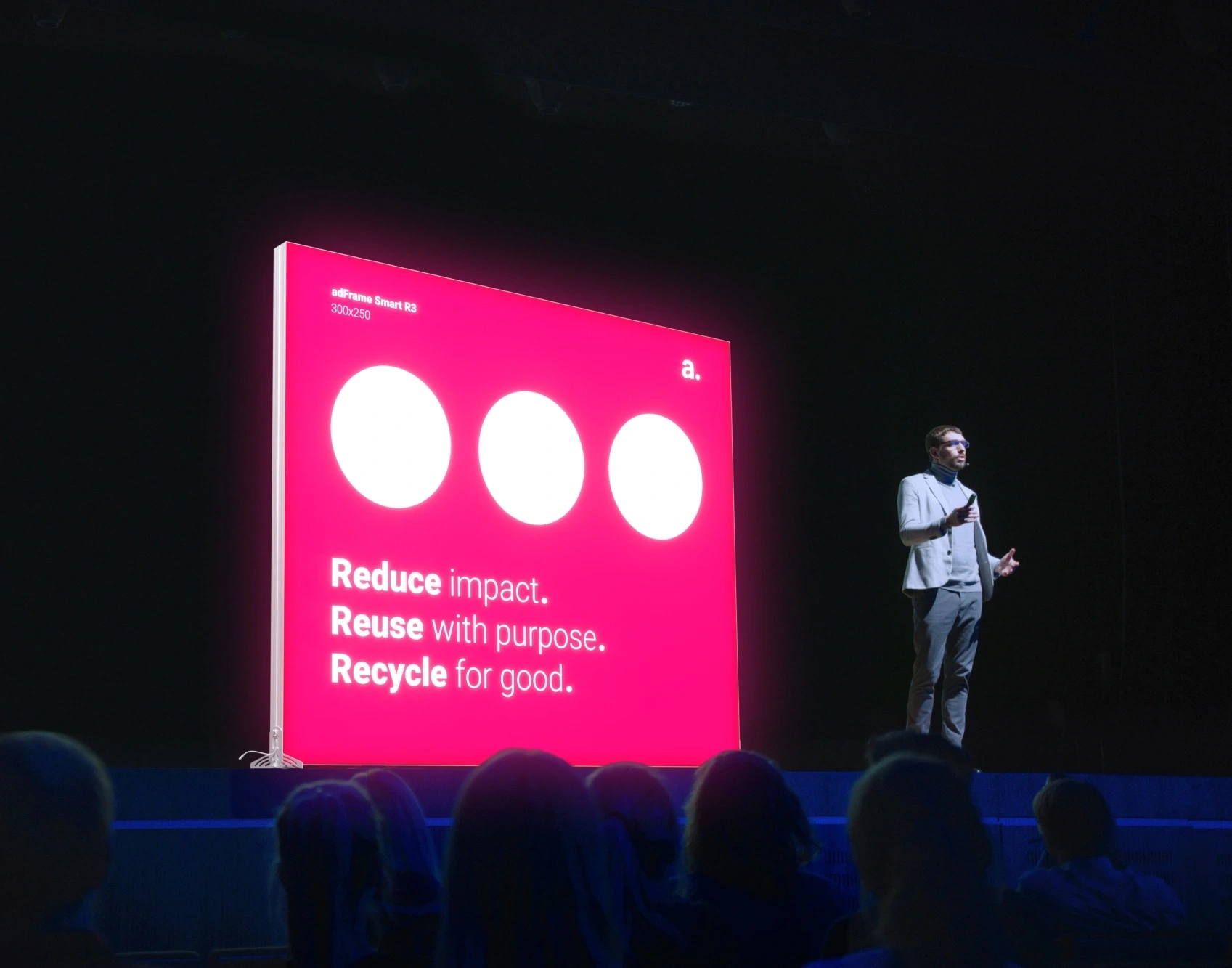 Ponente en un escenario junto a un gran panel publicitario LED de color fucsia con el mensaje "Reduce. Reuse. Recycle.", iluminado frente a una audiencia.