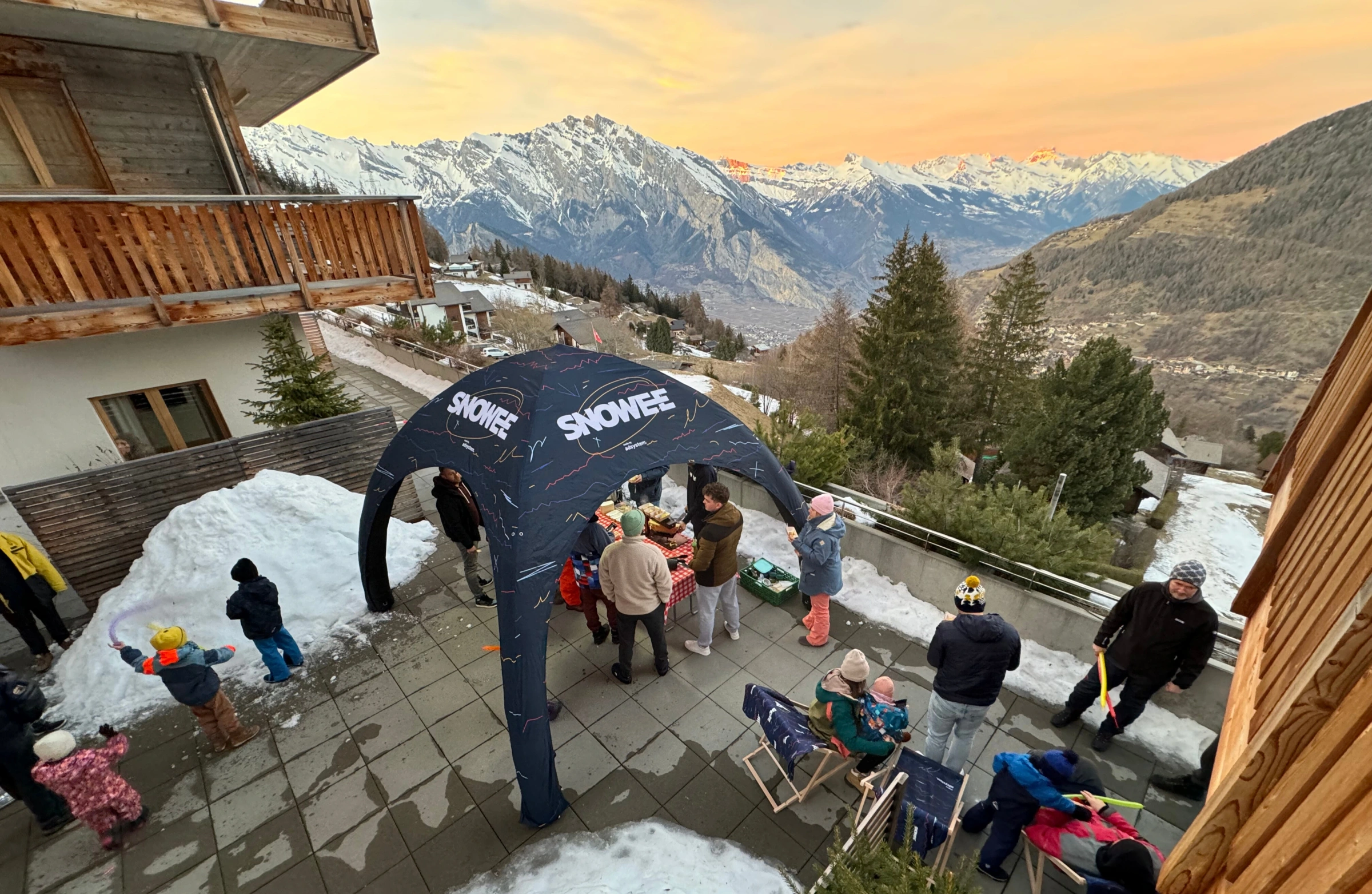 Grupa osób bawiących się i odpoczywających zimą na tarasie górskiego pensjonatu pod namiotem reklamowym z logo „SNOWEE”, z widokiem na ośnieżone Alpy o zachodzie słońca.