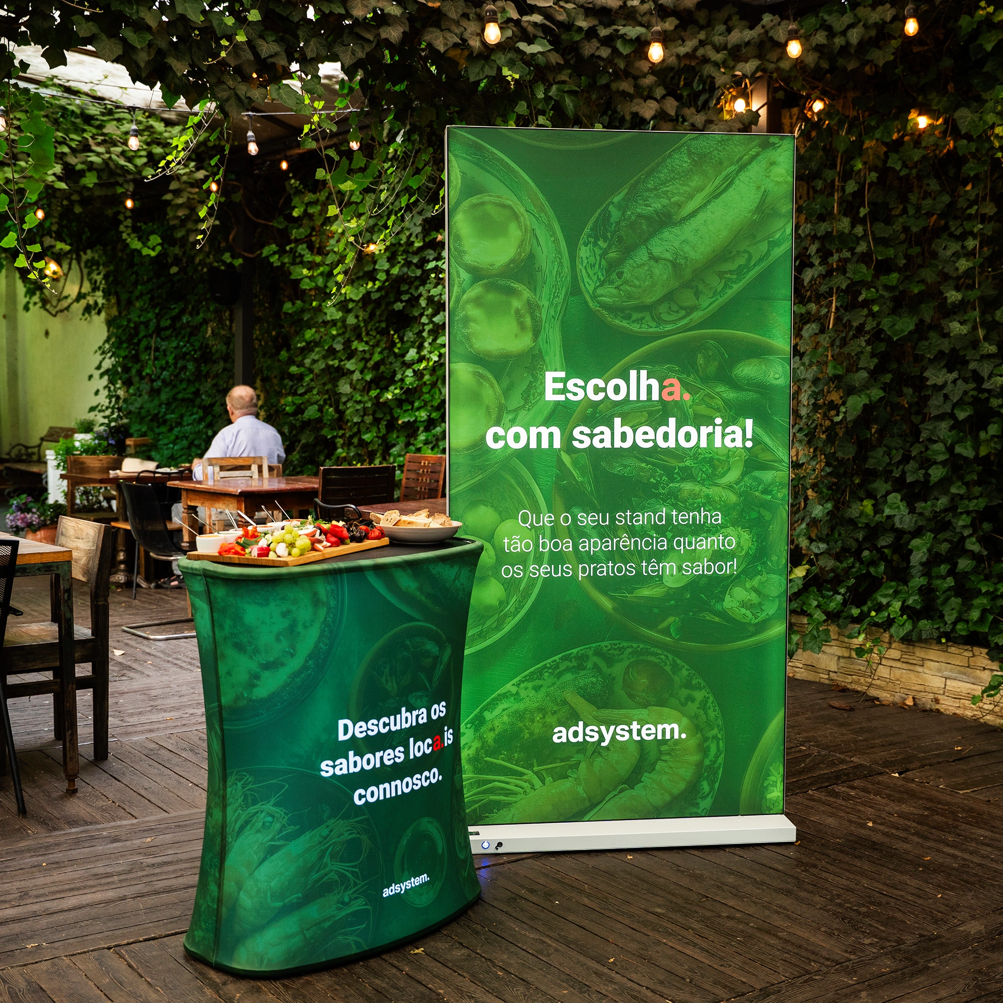 Trybunka i ścianka reklamowa adsystem w zielonej kolorystyce z portugalskimi hasłami, ustawione w ogrodowej przestrzeni eventowej pod girlandami świetlnymi.