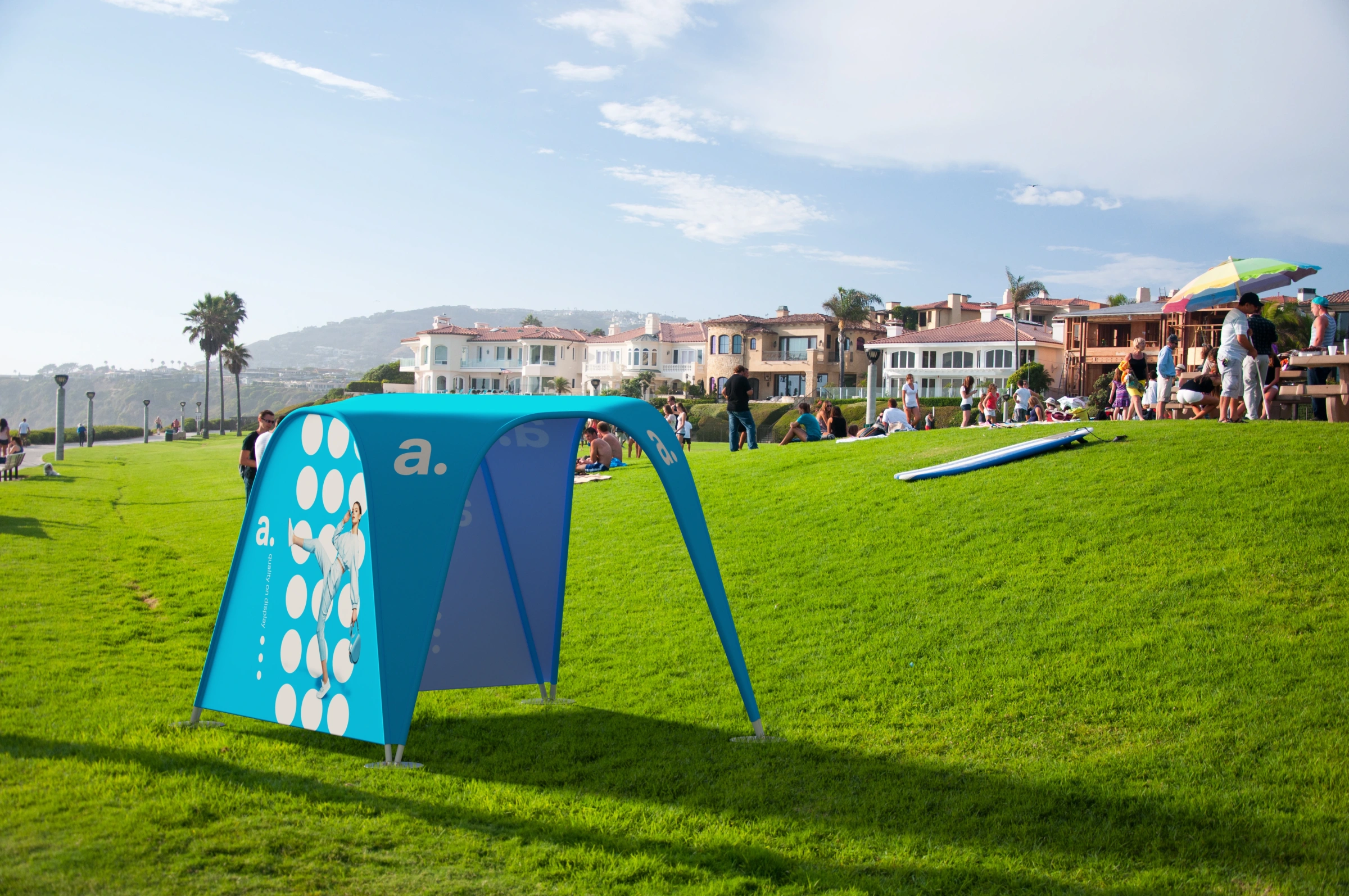 Namiot reklamowy adTent Vario 3x3 ustawiony na zielonym wzgórzu podczas imprezy plenerowej, z widokiem na luksusowe domy i uczestników wydarzenia w tle