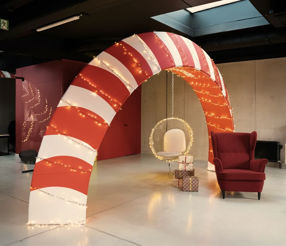 Świąteczna aranżacja z łukiem reklamowym w czerwono-białe pasy przypominające lukrowaną laskę cukrową, ozdobionym ciepłymi lampkami. Pod łukiem znajduje się przezroczysty wiszący fotel z podświetleniem i prezentami, a obok stoi czerwony fotel w nowoczesnym biurowym wnętrzu.