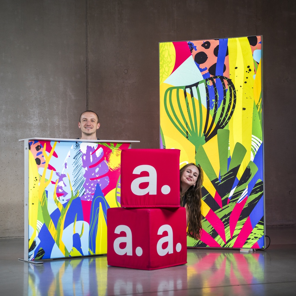 Dwie osoby pozujące przy podświetlanej trybunce, ściance SEG i pufach kostkach z wymiennymi pokrowcami, pokazując możliwości ponownego wykorzystania grafik.