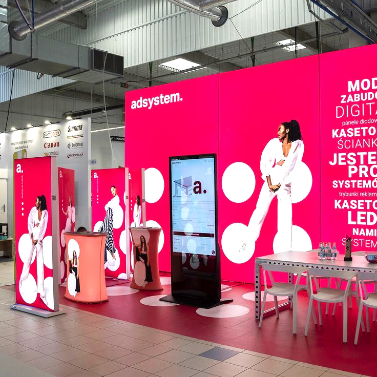 Stoisko targowe adsystem z podświetlaną zabudową w intensywnym różowym kolorze, ozdobioną dużymi białymi kropkami i zdjęciami modelek, wyposażone w trybunki reklamowe, ekran multimedialny oraz strefę spotkań z stołem i krzesłami.