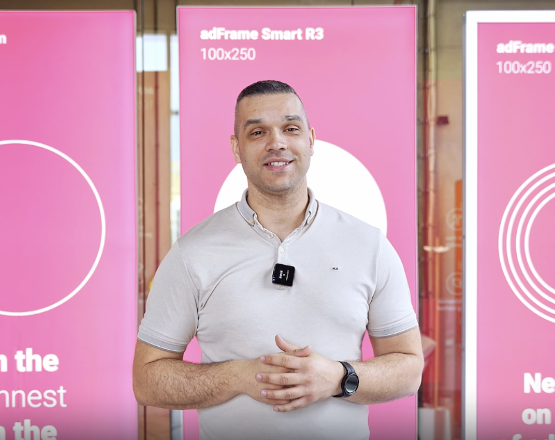 Hombre presentando los paneles publicitarios adFrame Smart R3 100x250 con fondo rosa durante una demostración en feria o showroom.